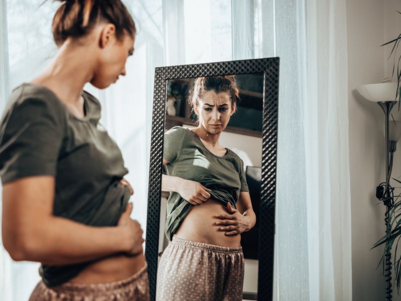 woman looks at herself in a stand up mirror and holds stomach to illustrate the problem of bloating or weight gain