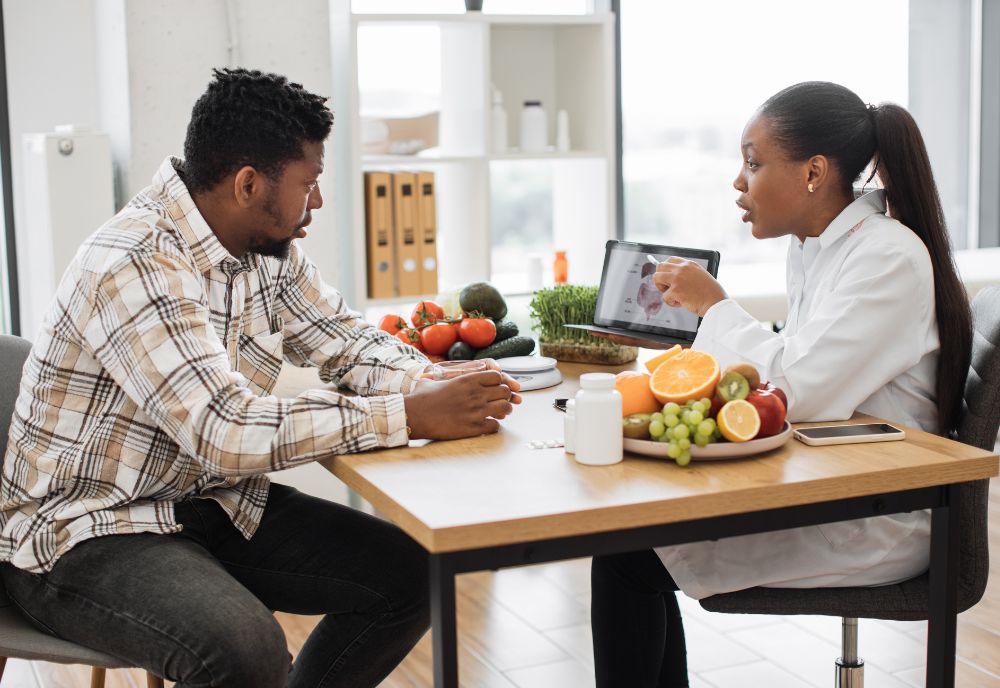 African american specialist in nutrition holding tablet with gastrointestinal tract on screen while counseling man at work. Focused woman explaining client about food intolerance during appointment.