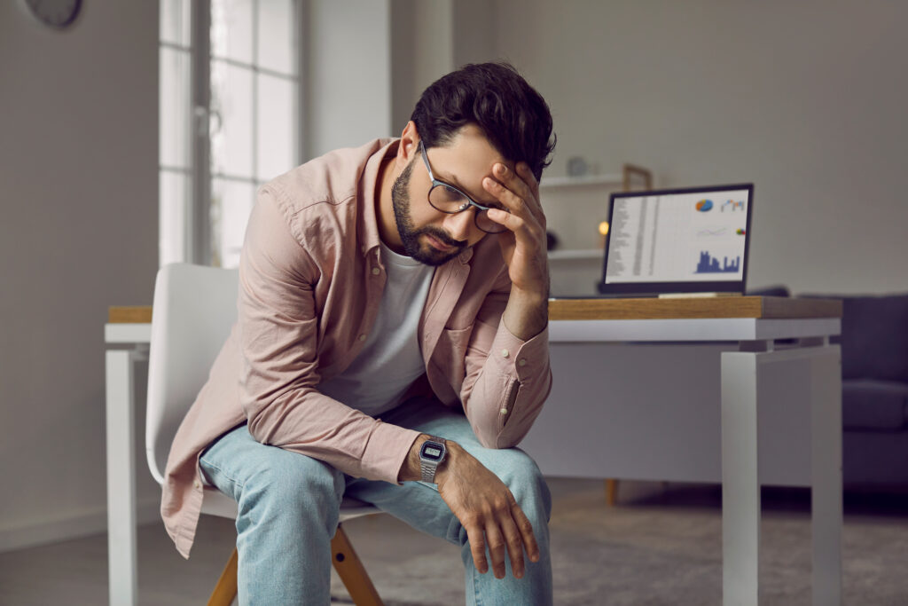 Stressed business man experiencing a burnout, sitting on a chair by his working desk with a laptop computer, holding his hand on his head, feeling tired and frustrated. Stress and burnout concept