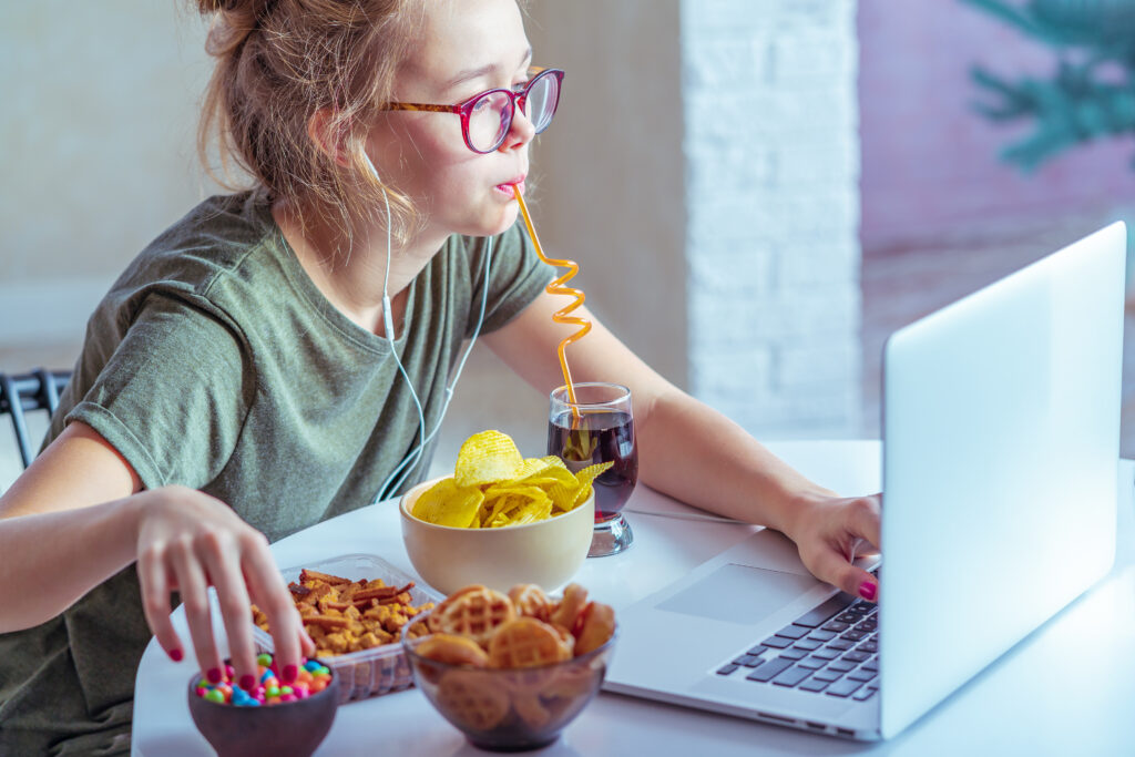 Girl works at a computer and stress eats fast food. Unhealthy food: chips, crackers, candy, waffles, cola. Junk food, concept.