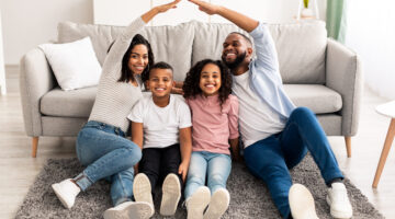 Family Care, Protection And Insurance Concept. Portrait of smiling African American parents making symbolic roof of hands above their happy children, sitting on the floor carpet in living room at home