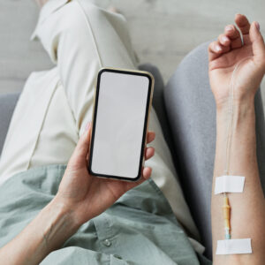 Minimal top view close up of unrecognizable woman getting IV drip and using smartphone with blank screen, copy space with a person getting an IV therapy