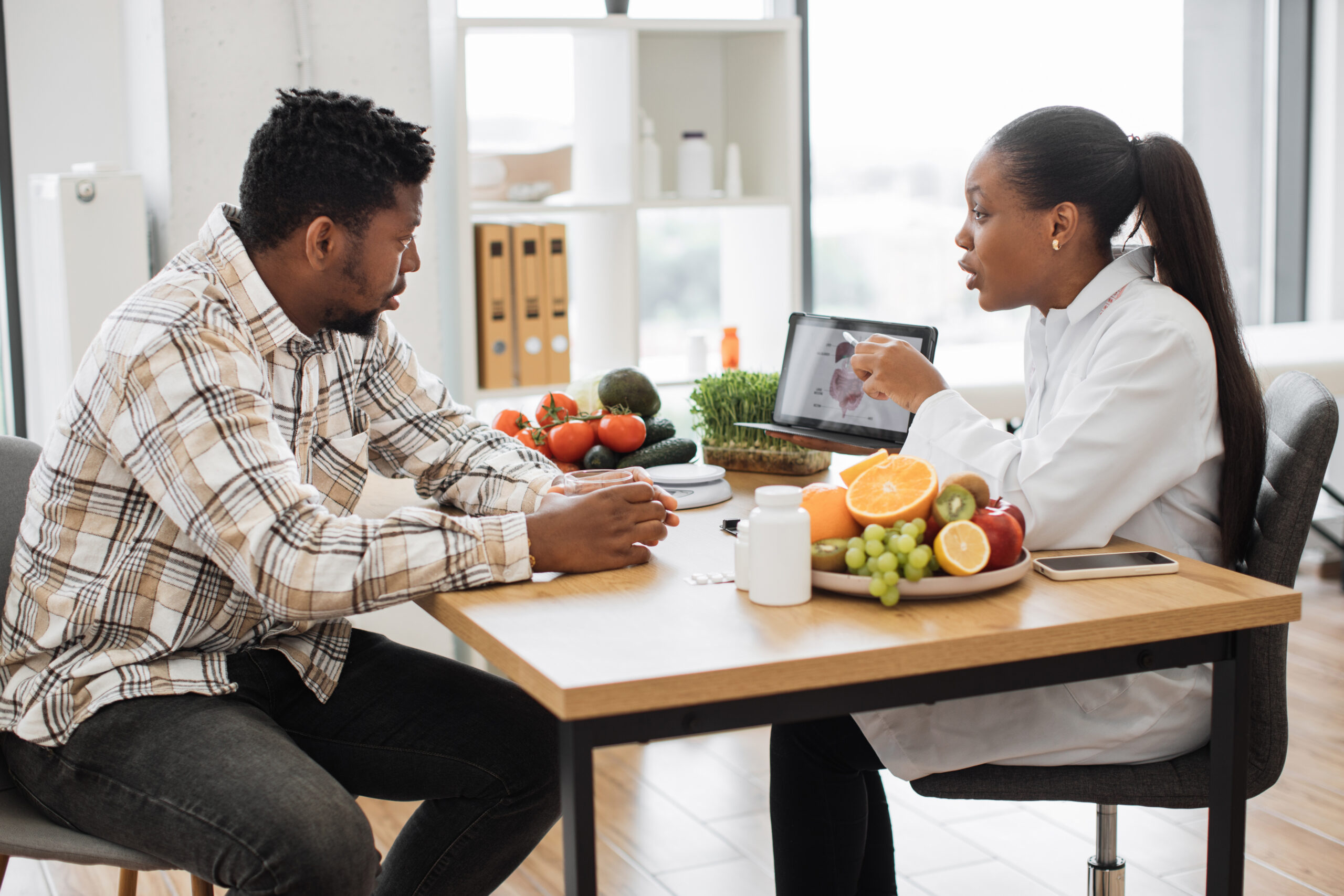 African american specialist in nutrition holding tablet with gastrointestinal tract on screen while counseling man at work. Focused woman explaining client about food intolerance during appointment.