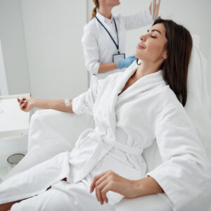 Side view portrait of attractive woman in white bathrobe sitting in armchair with closed eyes while receiving IV infusion