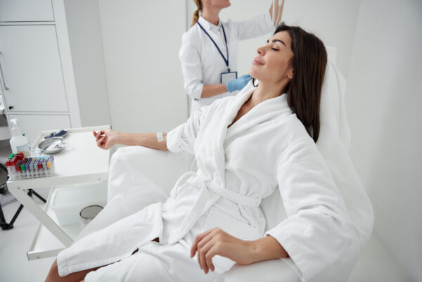 Side view portrait of attractive woman in white bathrobe sitting in armchair with closed eyes while receiving IV infusion