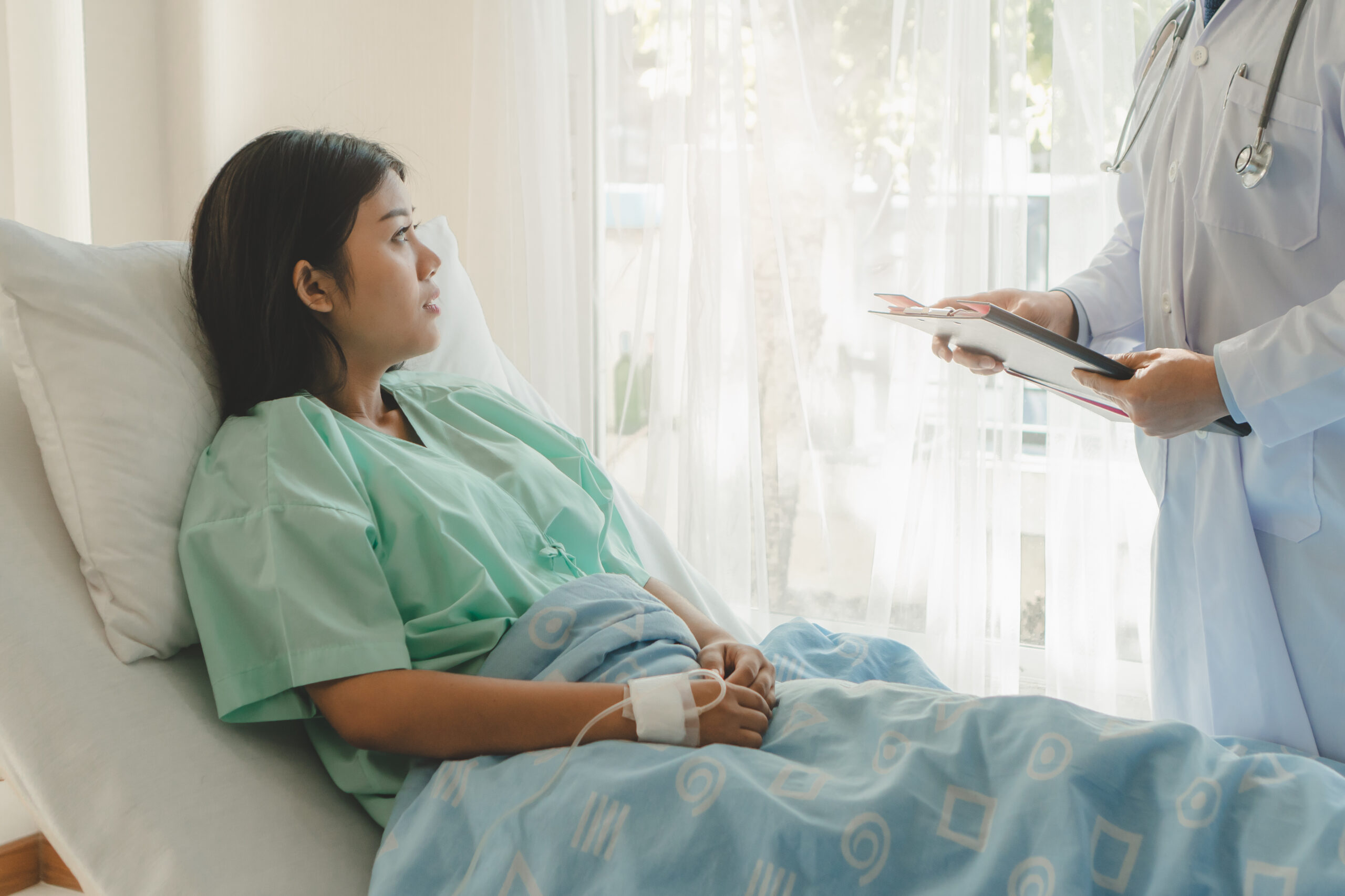 Licensed young woman sitting on the bed and looking outside window in hospital worry about her illness.