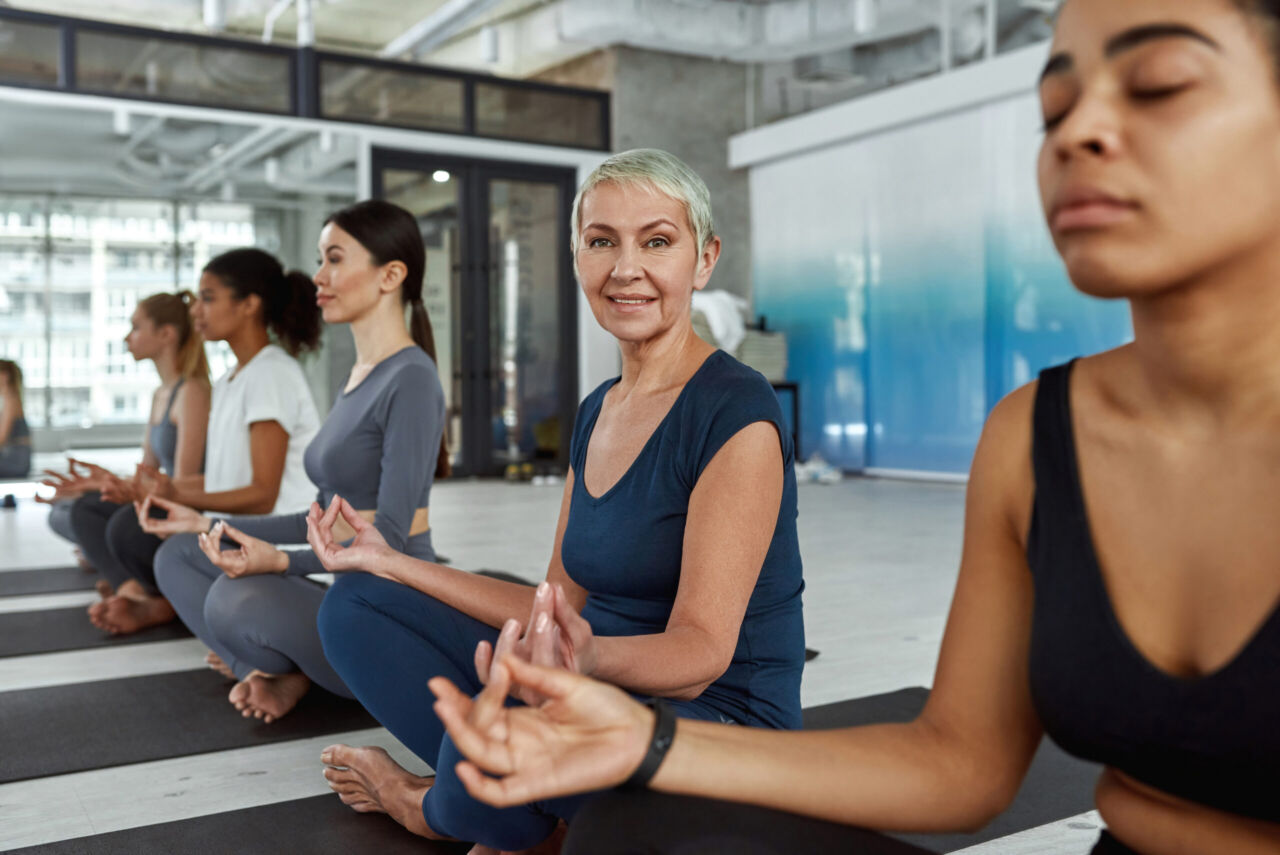 Smiling sporty Caucasian middle-aged woman in sportswear practice yoga meditate at group training in club. Happy active do sports exercise work out at session. Healthy lifestyle concept.